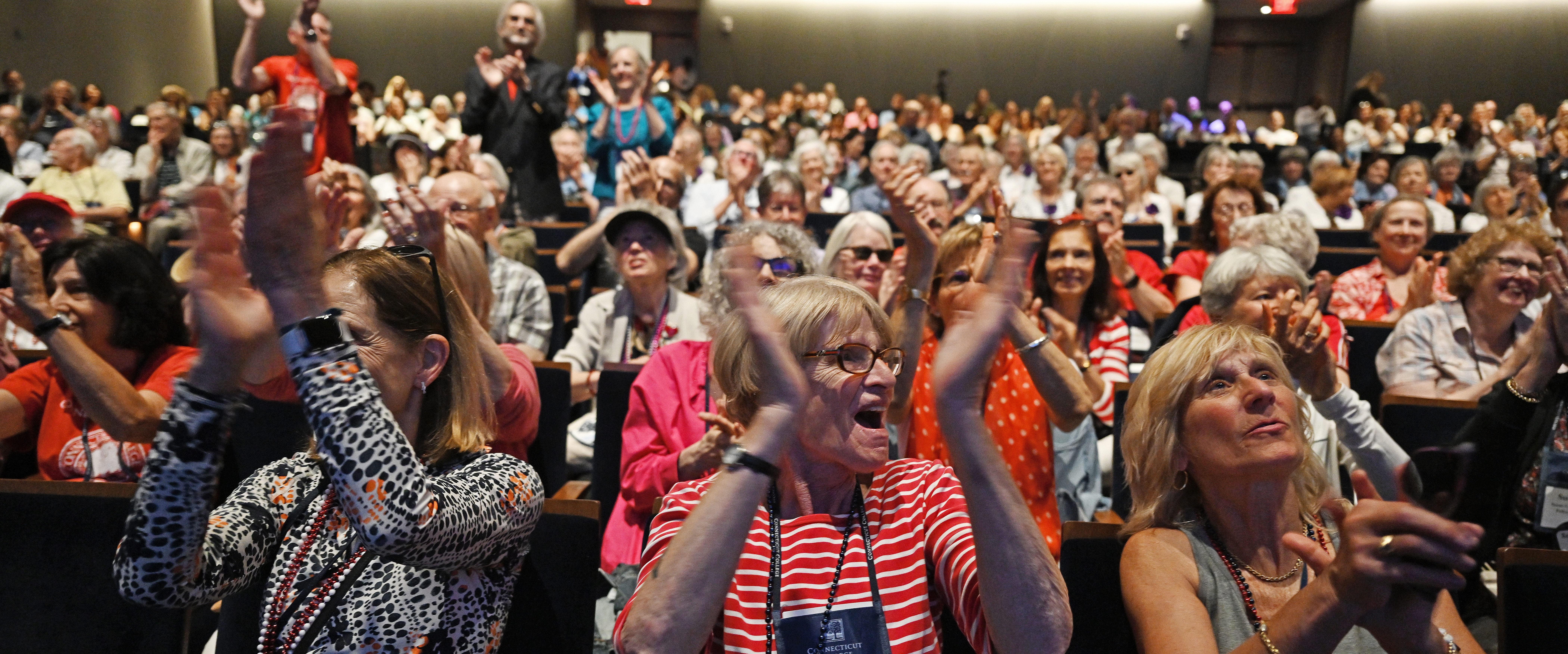 A crowd of college alumni gathered in a theater space applaud and cheer