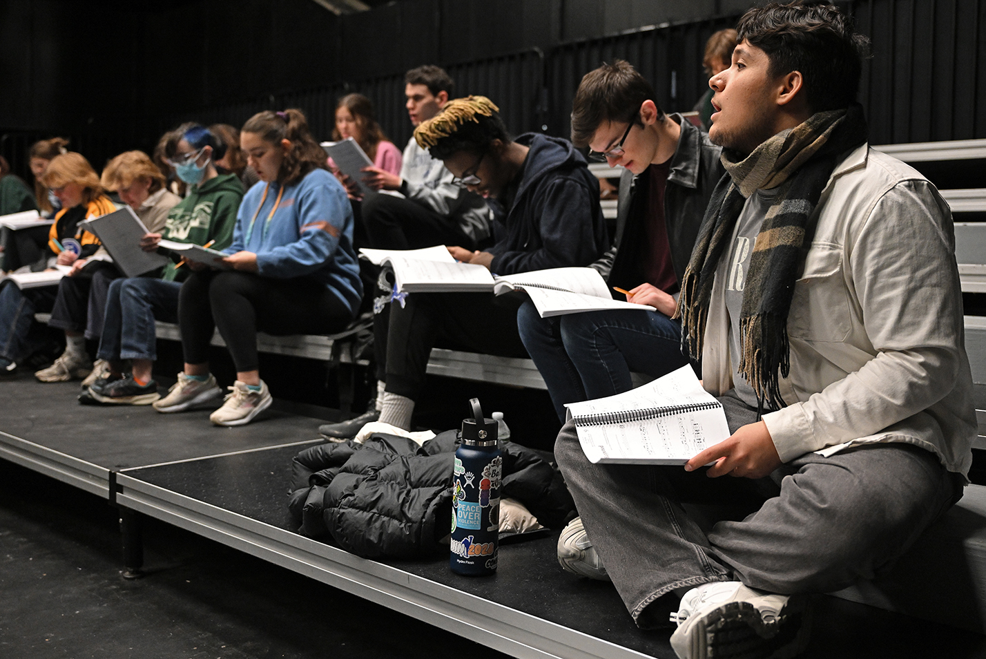 A group of seated actors practice a musical number during rehearsal in a black box theater space.